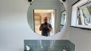 A bathroom with a round mirror, a white sink, a wooden vanity, a towel rack, and a window with a view of trees. - Quiet, private Cosy Cabin with en-suite. Access to bike paths, river and pub. (Puketapu)