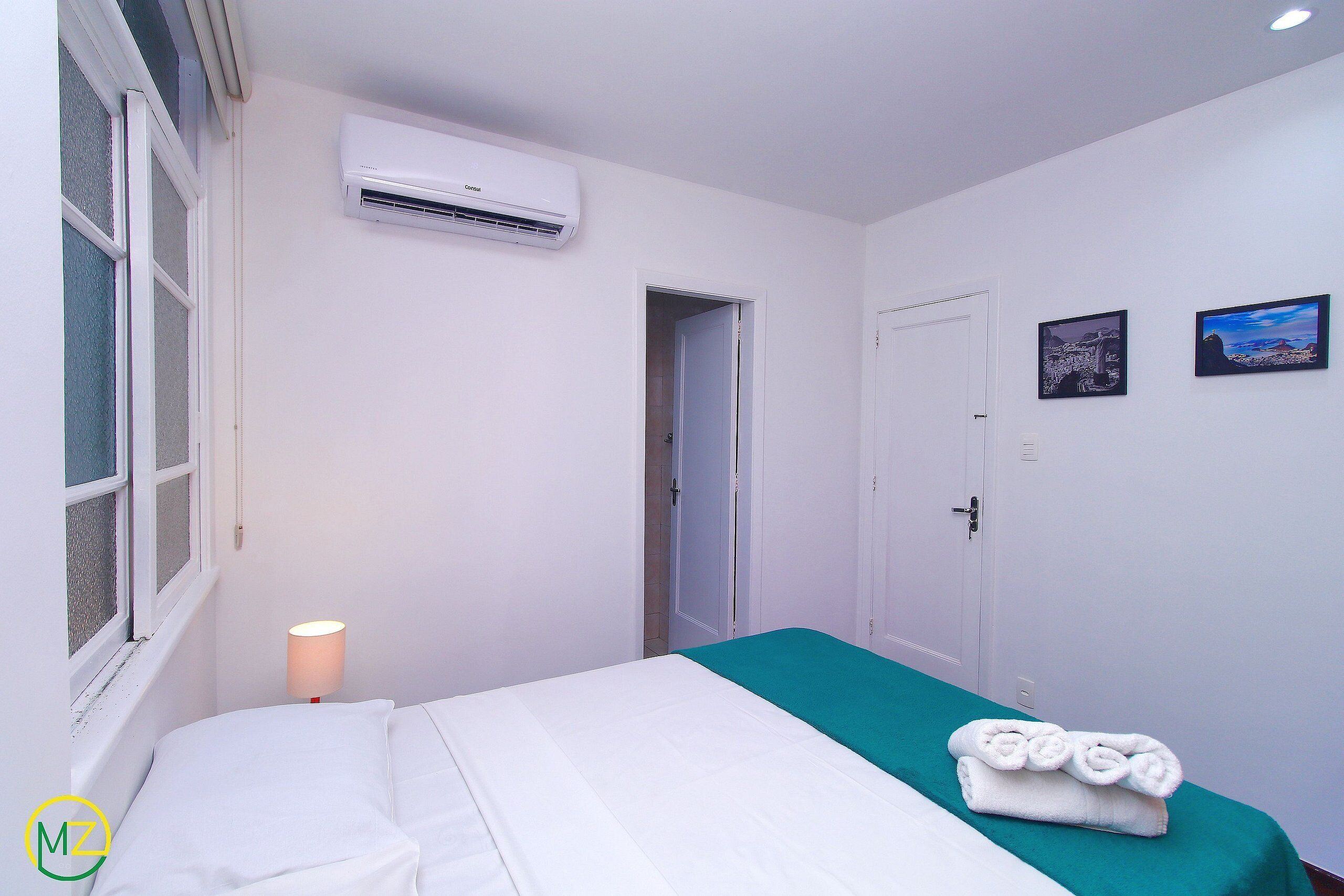 A hotel room with a bed, a window, an air conditioner, a door, and two framed pictures on the wall.