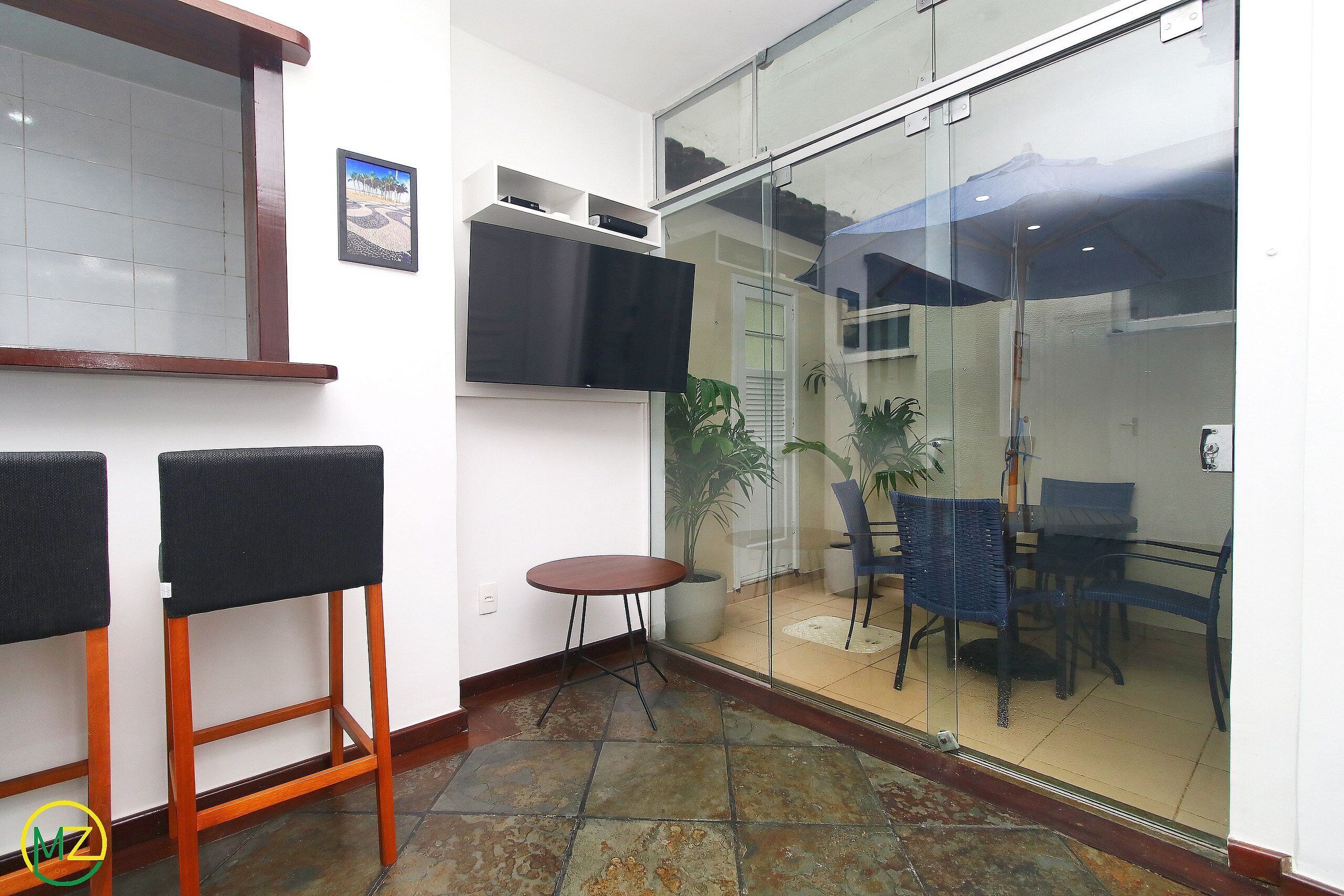 A small, enclosed patio area with a glass door leading to a balcony. Inside, there are two wooden chairs, a small round table, and a mounted flat-screen TV.