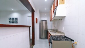 Eine schmale Küche mit weißen Fliesen, einem Edelstahlkühlschrank, einem Gasherd und Holzschränken.