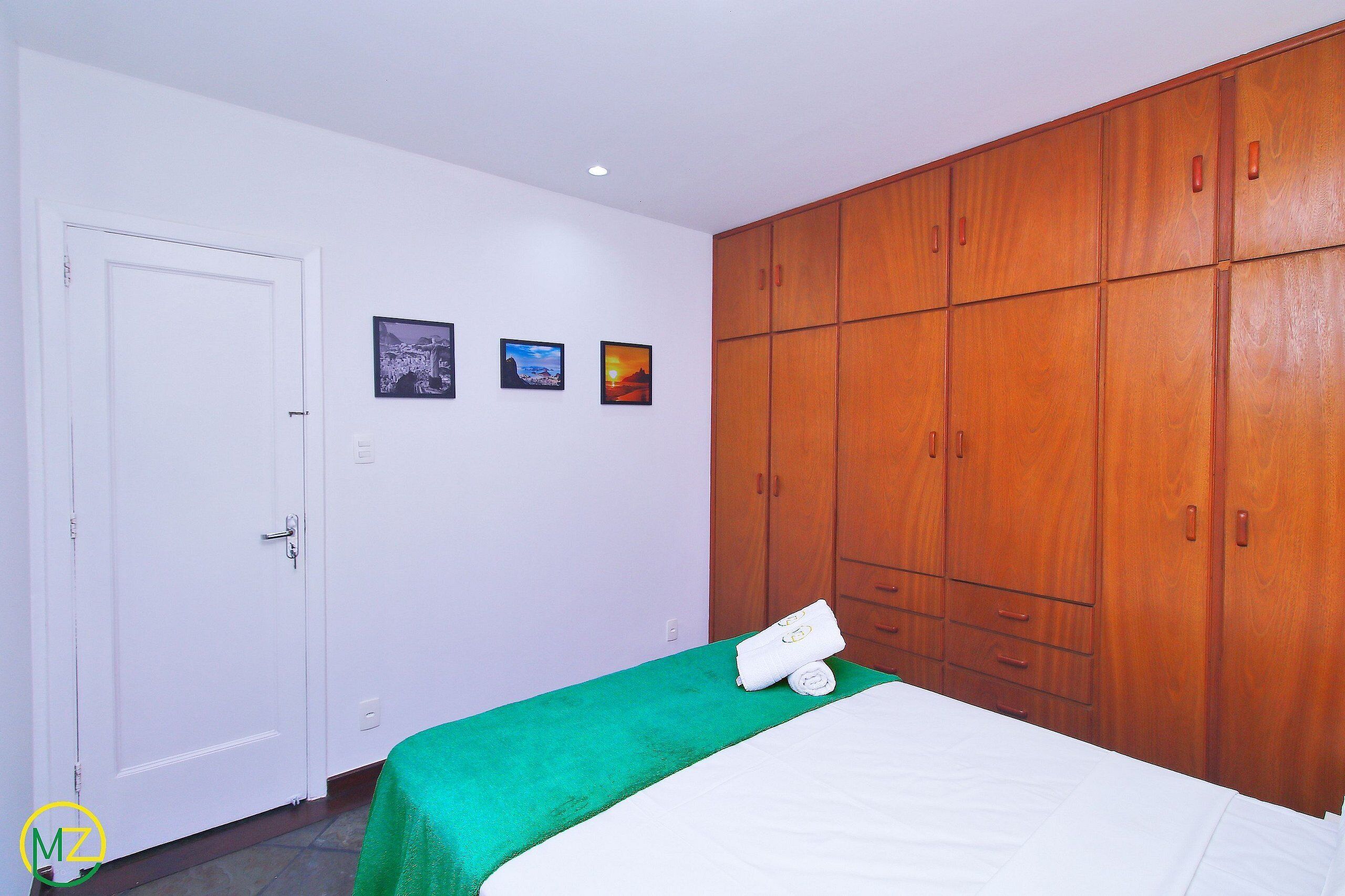 A hotel room with a bed, a green bedspread, a wooden wardrobe, and three framed pictures on the wall.