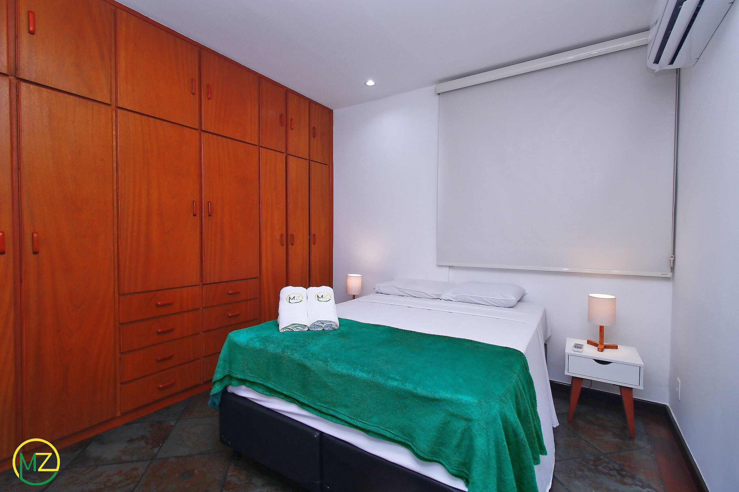 A bedroom with a bed, a green blanket, a nightstand with a lamp, and wooden wardrobe.