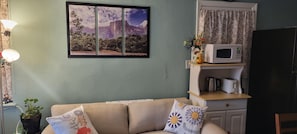 Olohuoneessa on beige sohva, seinälle asennettu televisio, mikroaaltouuni ja pieni keittiöalue.