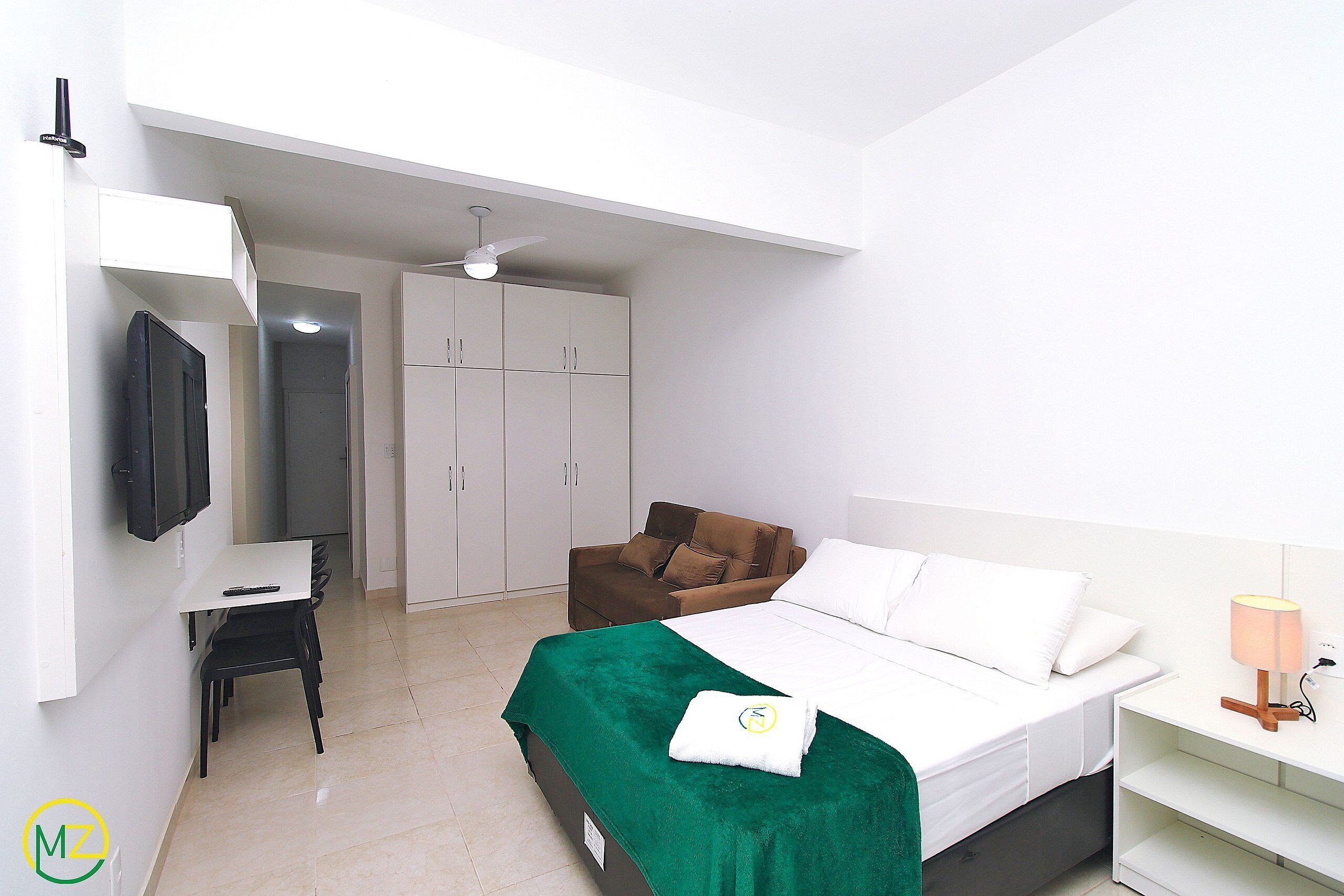 A hotel room with a bed, a desk, a chair, a TV, a wardrobe, and a ceiling fan.