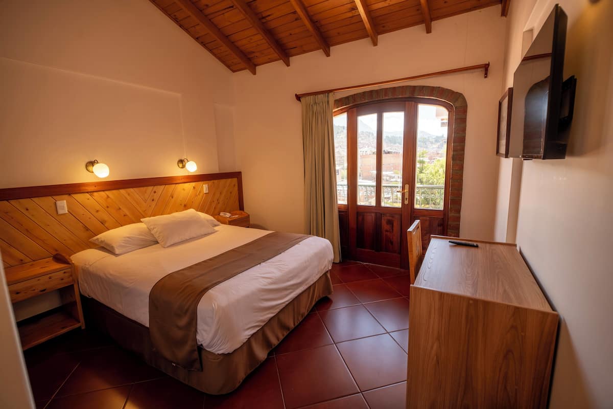 Un dormitorio con cabecera de madera, cama con ropa blanca, mesita de noche, escritorio de madera y televisor montado en la pared.