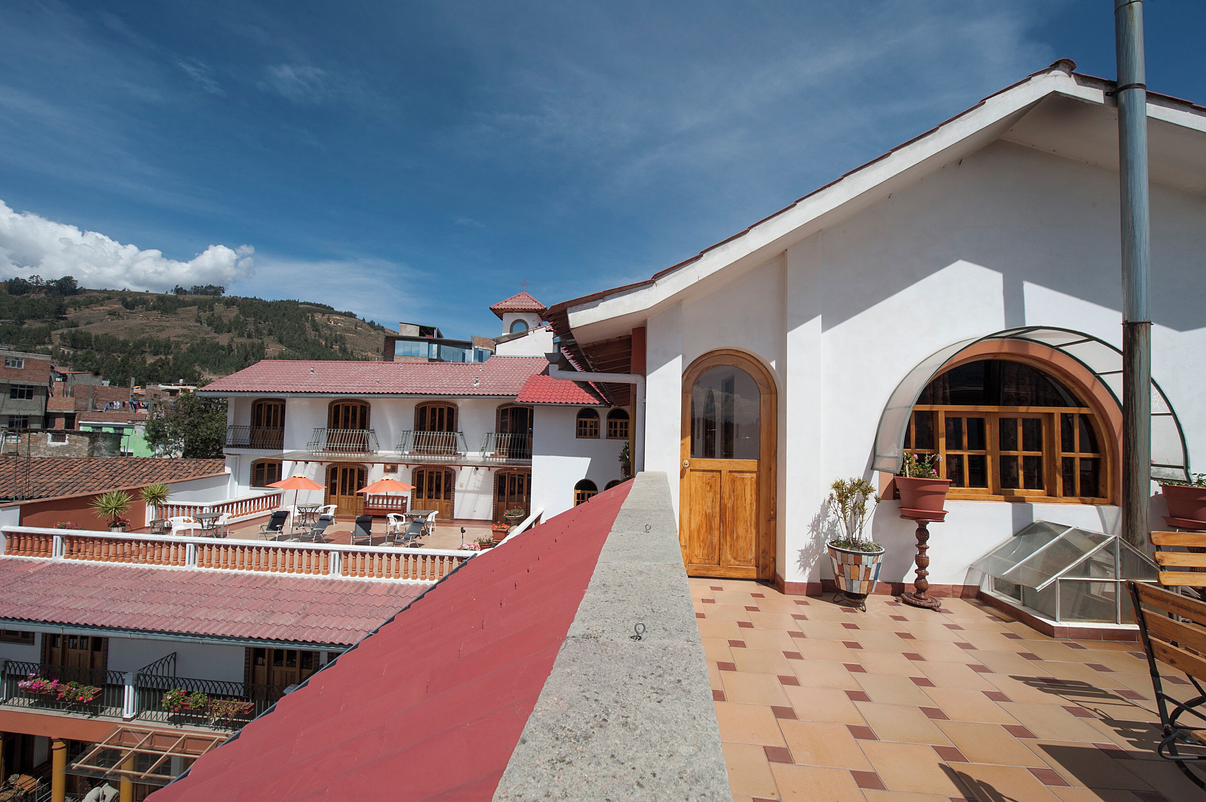 Um edifício de dois andares com telhado vermelho, varanda com mobiliário exterior e uma grande porta de madeira.