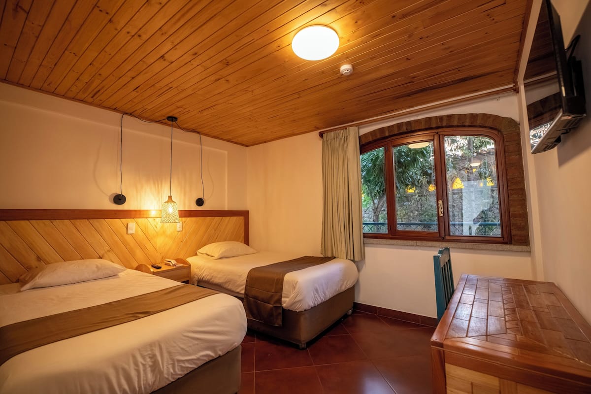 Una habitación con dos camas, techo de madera, ventana con cortinas y una mesa.