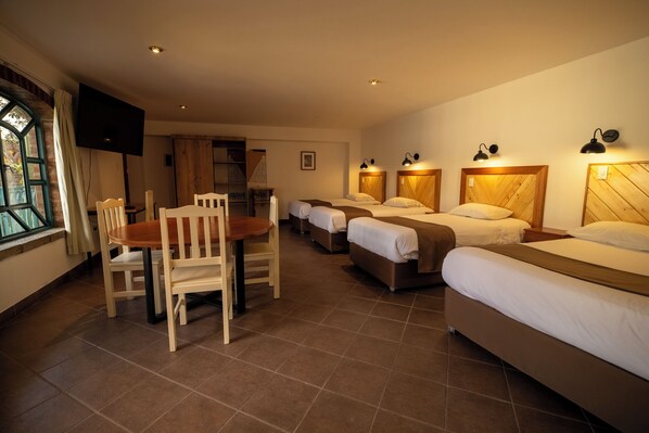 Ein Hotelzimmer mit vier Einzelbetten, einem Holzesstisch mit Stühlen, einem an der Wand befestigten Fernseher und einem Fenster mit Vorhängen.