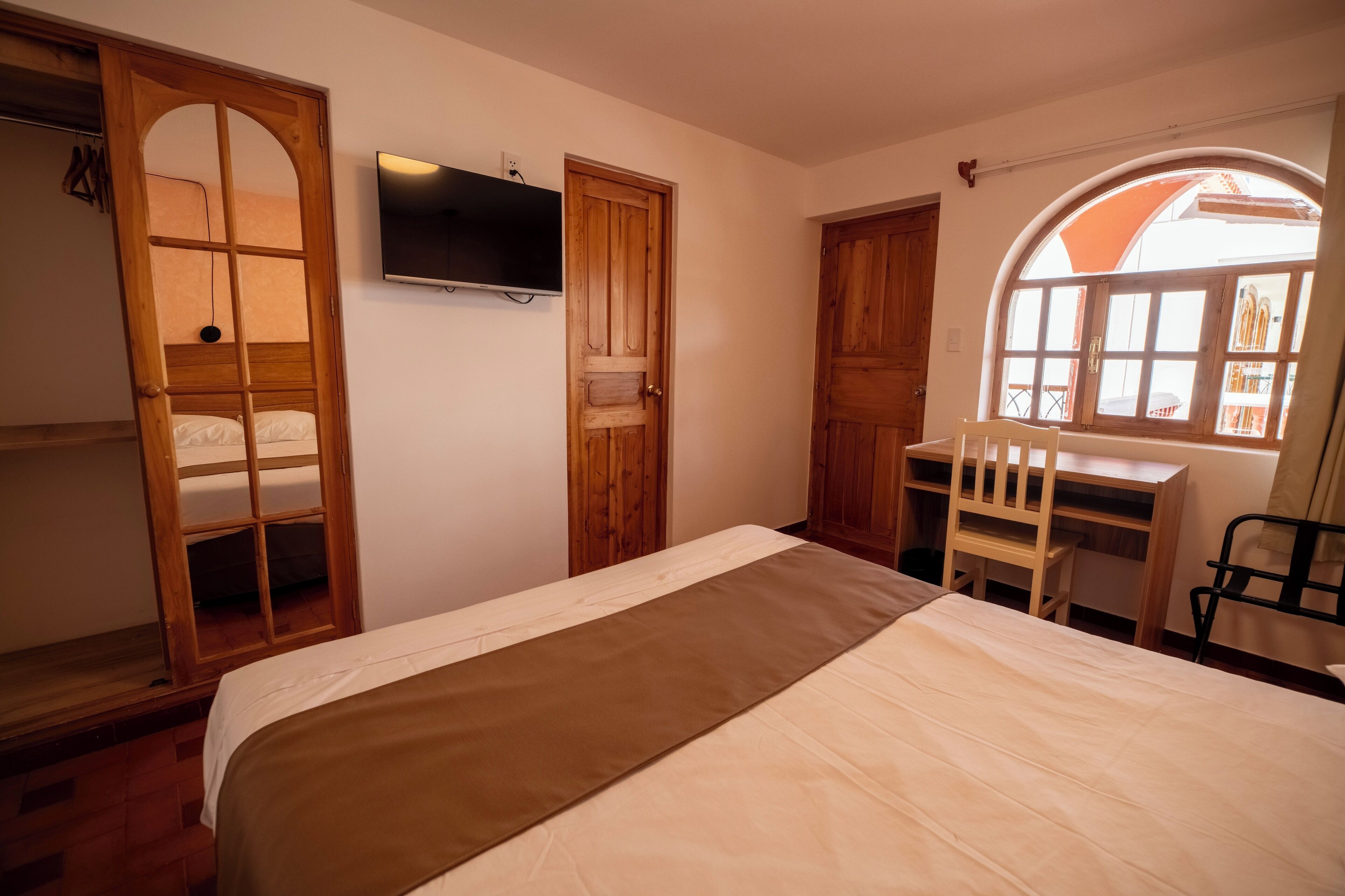 A bedroom with a bed, wooden wardrobe, desk, chair, and a TV.