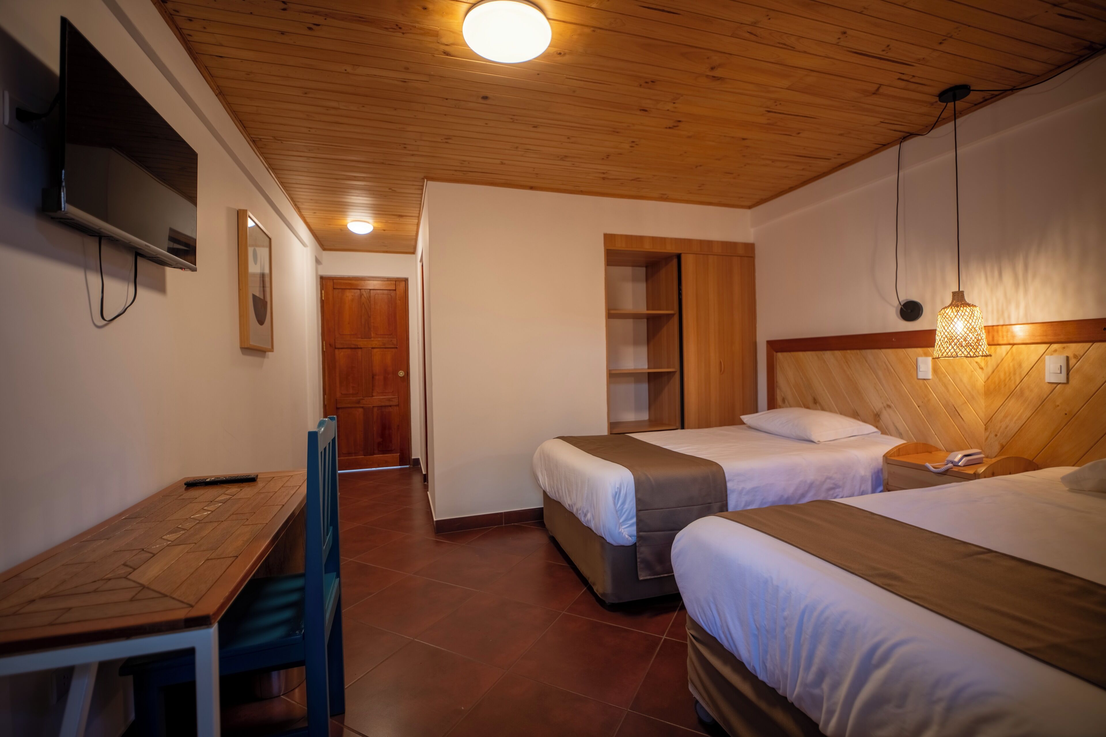 A hotel room with two beds, a wooden desk, a television, and a wooden ceiling.