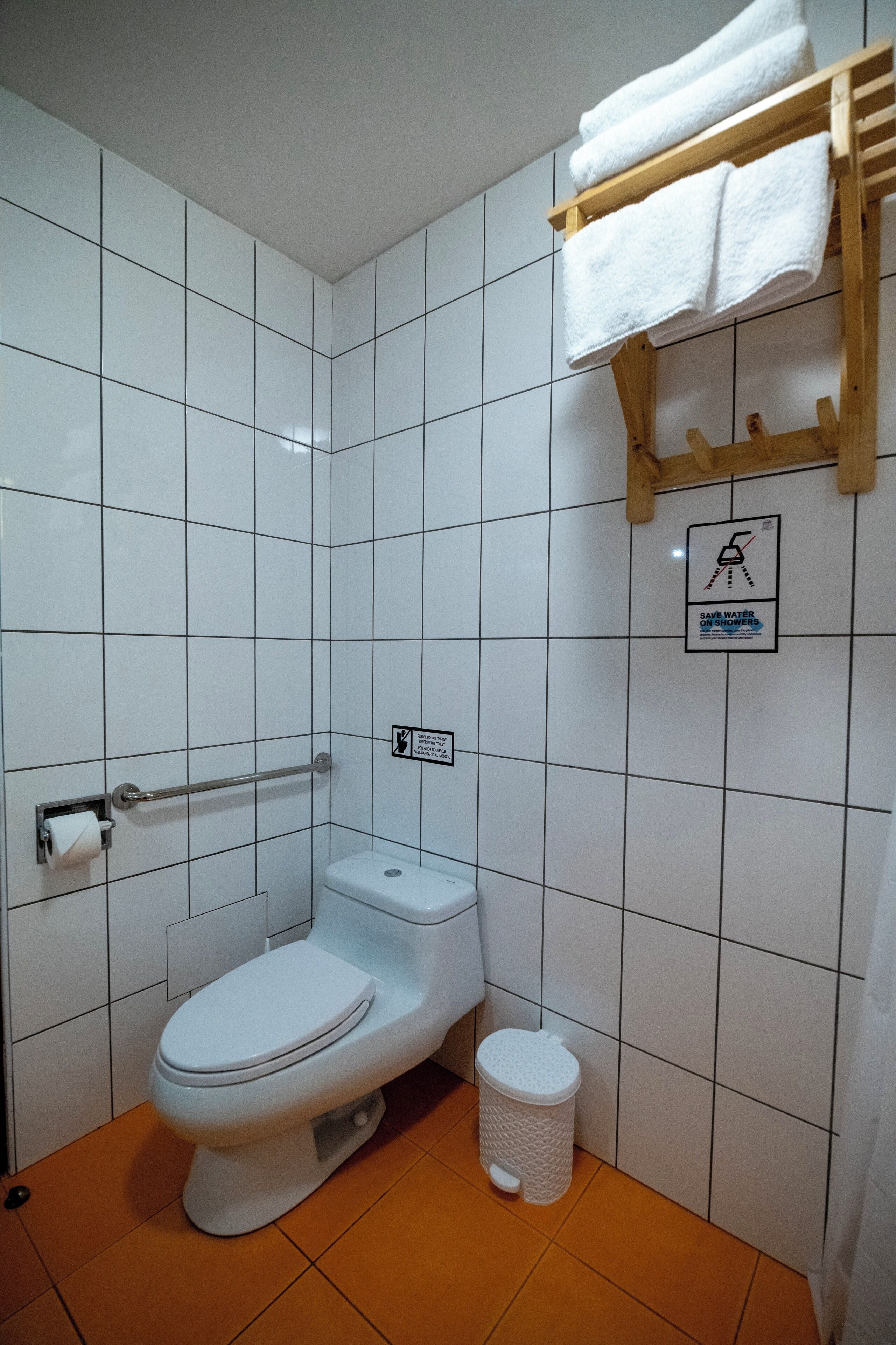 Un baño pequeño y limpio con azulejos blancos, un inodoro, un secador de manos, un cubo de basura y un perchero para toallas.