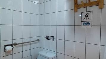 Un baño pequeño y limpio con azulejos blancos, un inodoro, un secador de manos, un cubo de basura y un perchero para toallas.