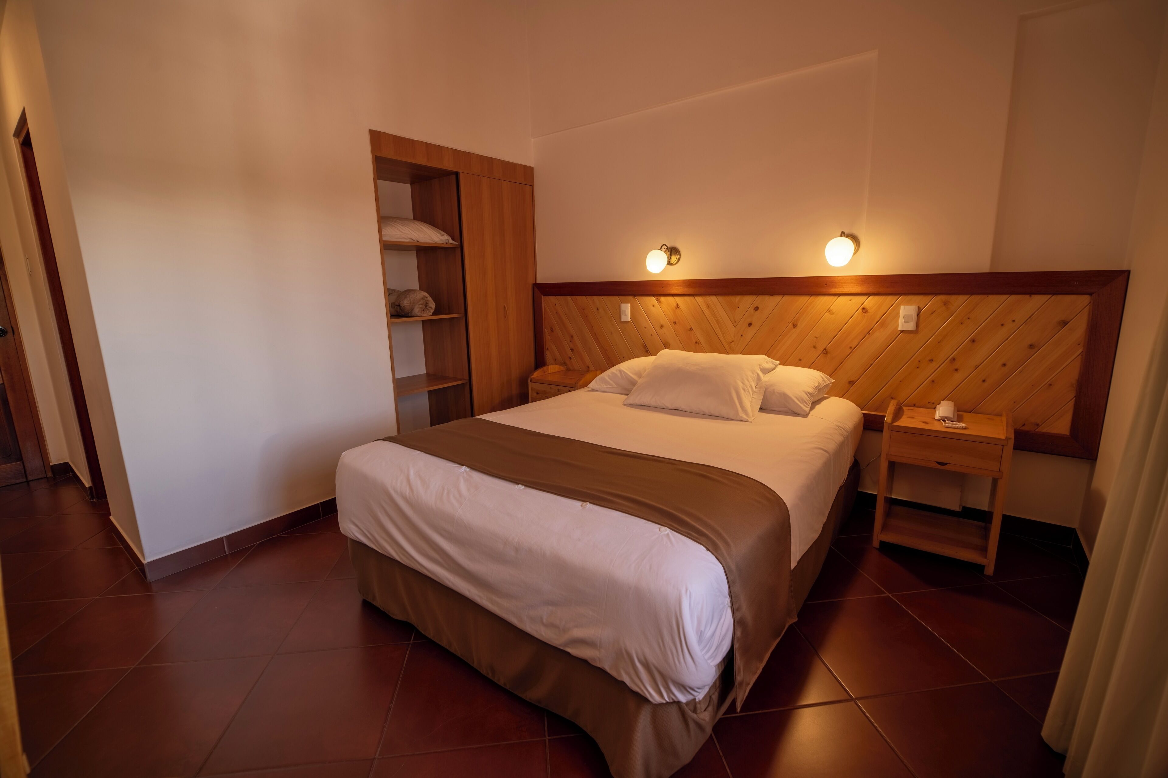 Una cama bien hecha con cabecera de madera, una mesita de noche con lámpara y una estantería a la izquierda.