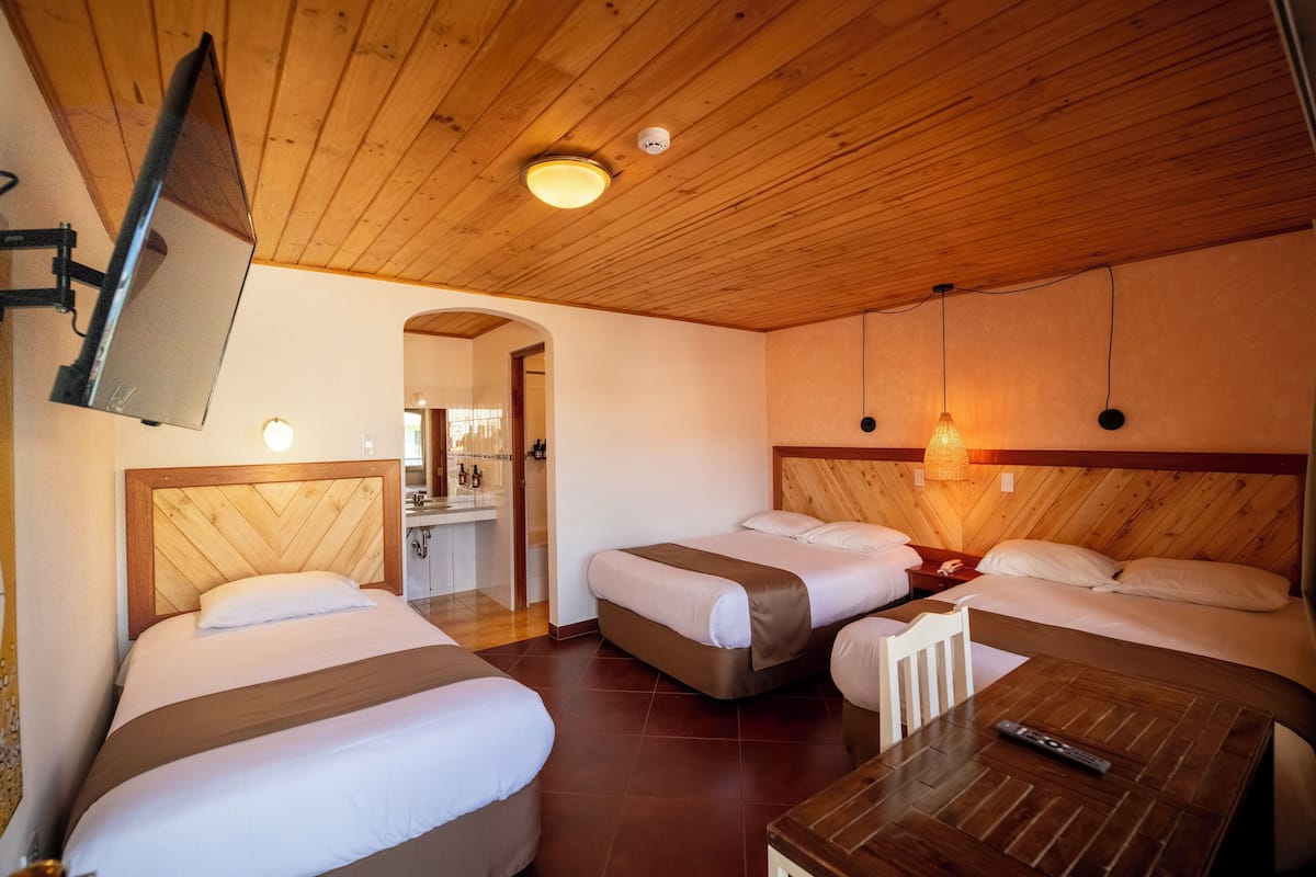 Habitación de hotel con tres camas, techo de madera, televisor montado en la pared y una pequeña mesa con un control remoto.