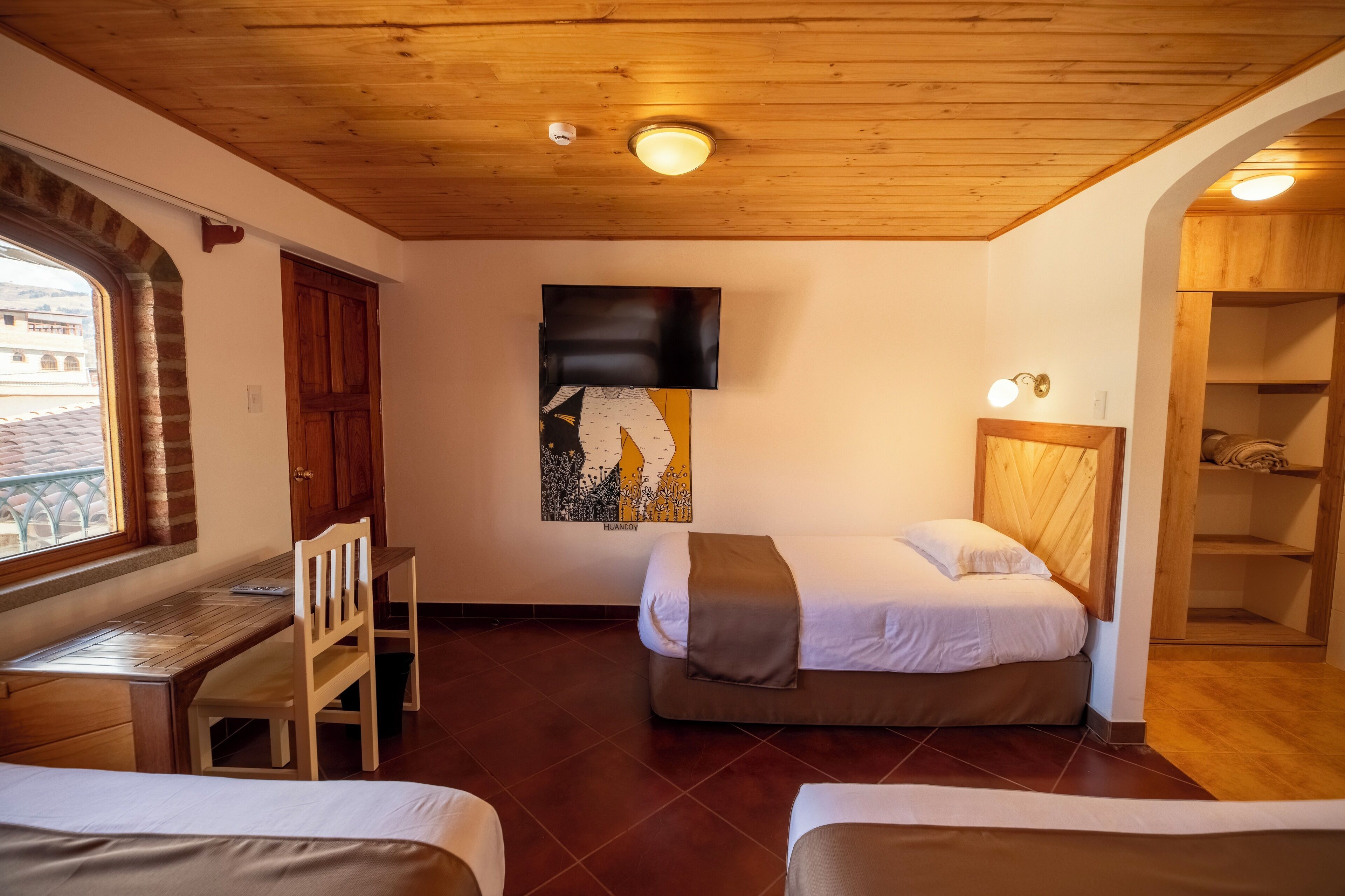 Una habitación con techo de madera, una cama con cabecera, un escritorio con silla, un televisor y una ventana con vistas a edificios.