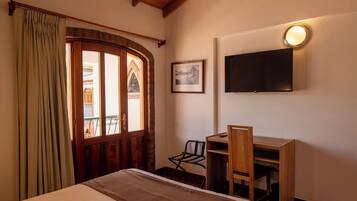 A hotel room with a wooden ceiling, a bed, a desk with a chair, a TV mounted on the wall, and a window with curtains.