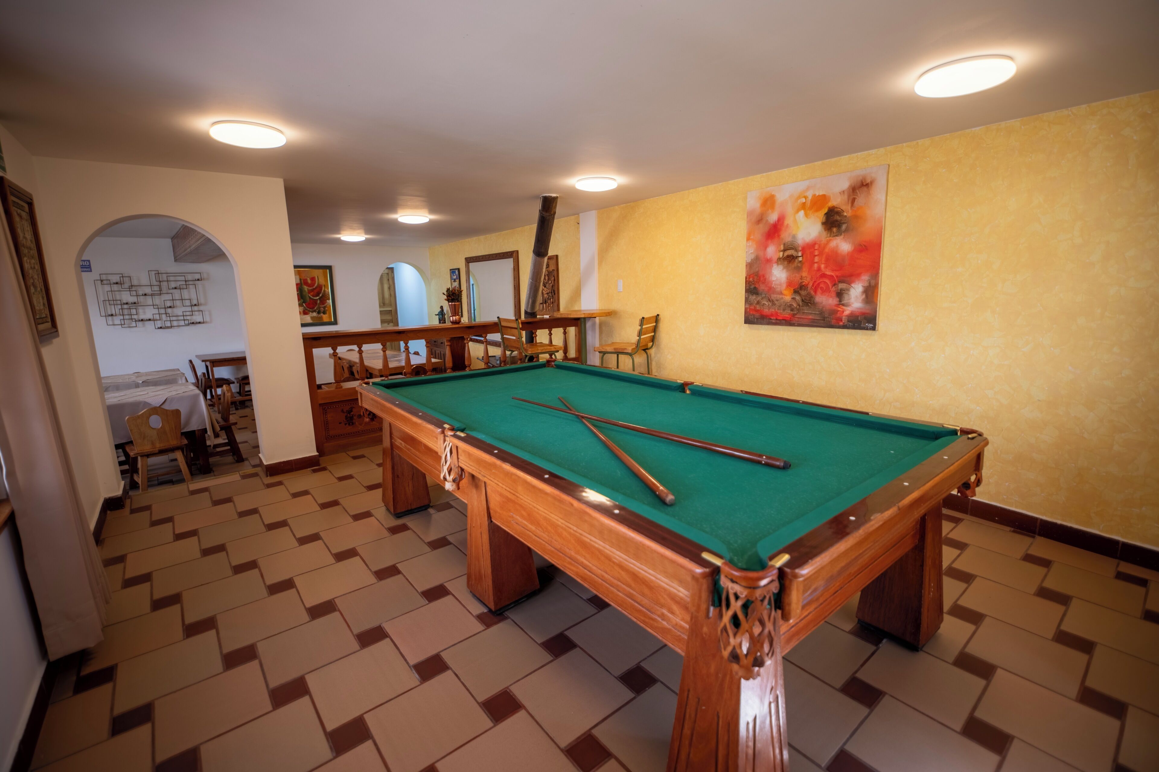 A pool table in a room with a herringbone floor pattern, a dining area with chairs, and a painting on the wall.