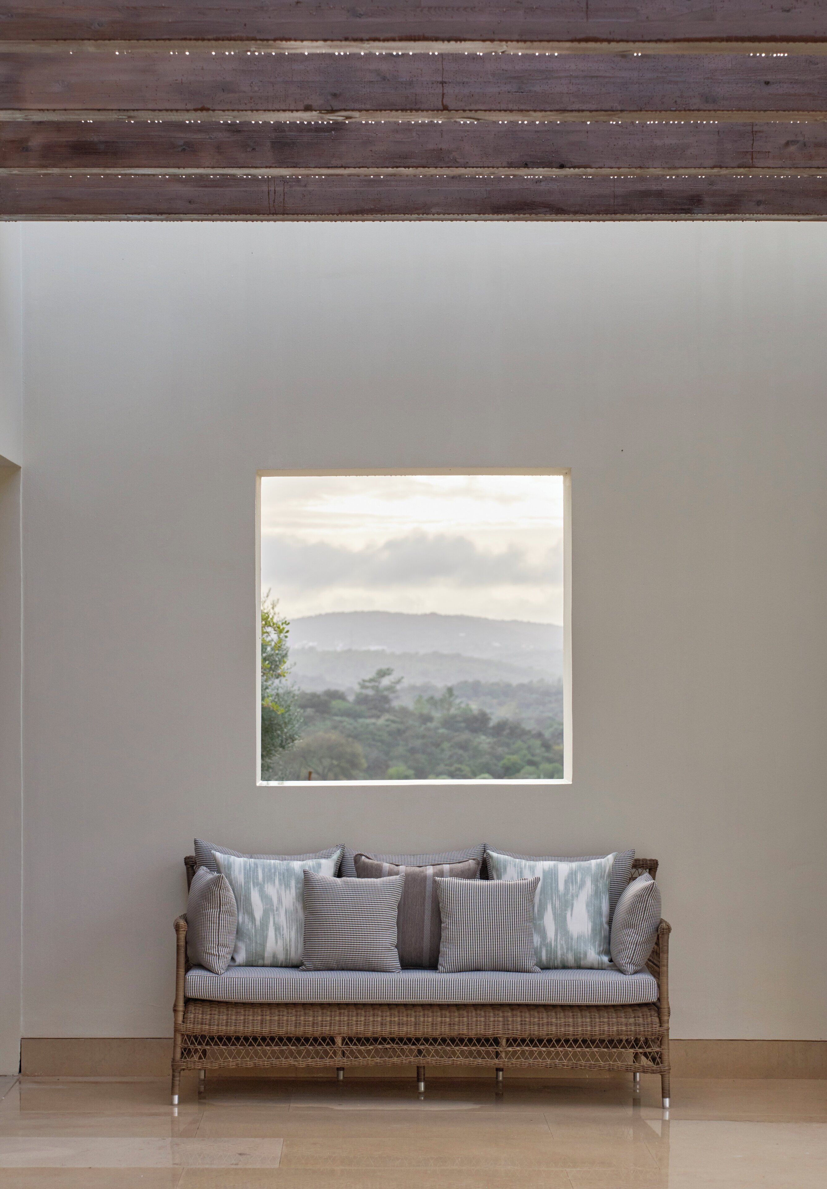A room with a large window, a wicker sofa with patterned cushions, and a plain wall.