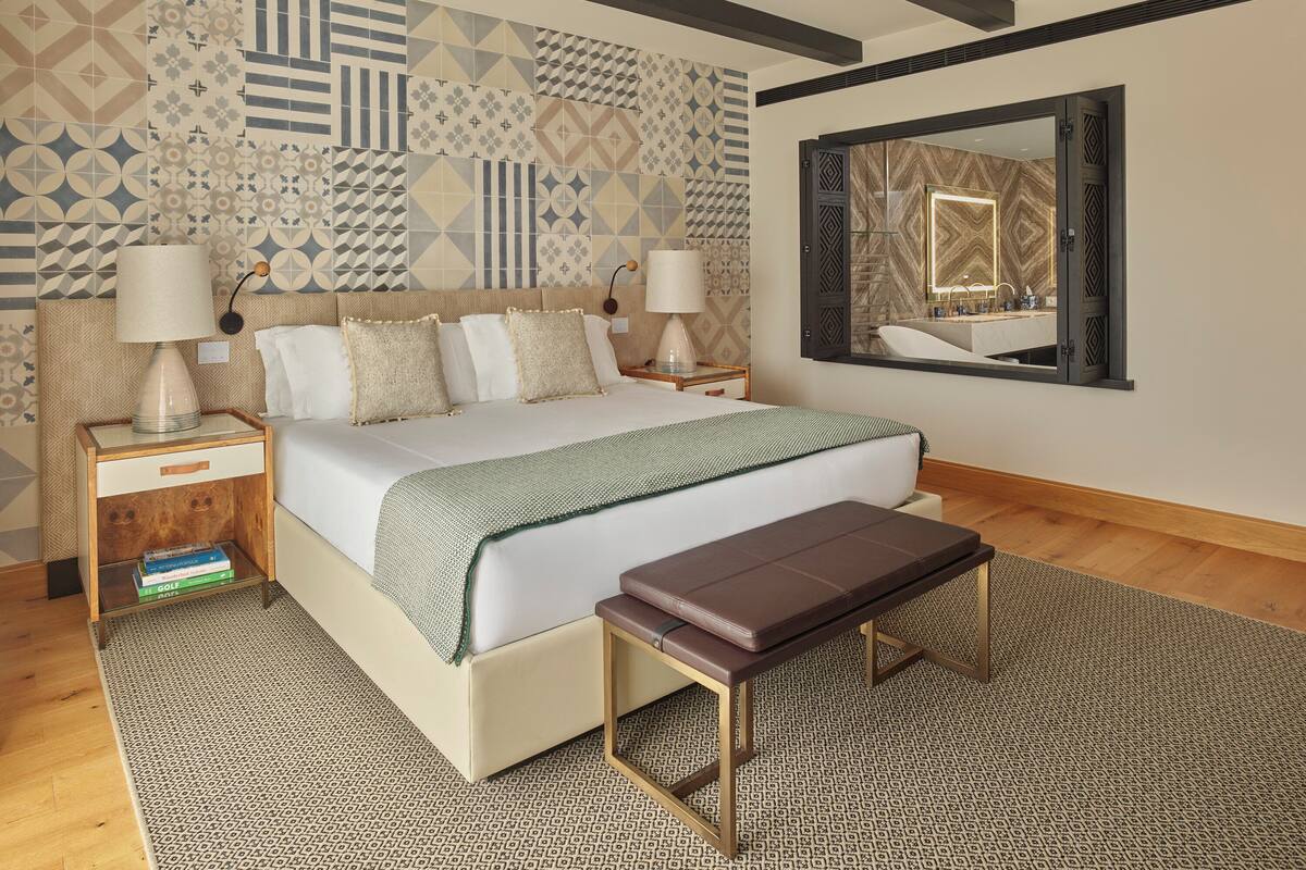 A modern hotel room with a large bed, two bedside tables with lamps, a bench, a patterned rug, and a decorative wall with geometric designs.