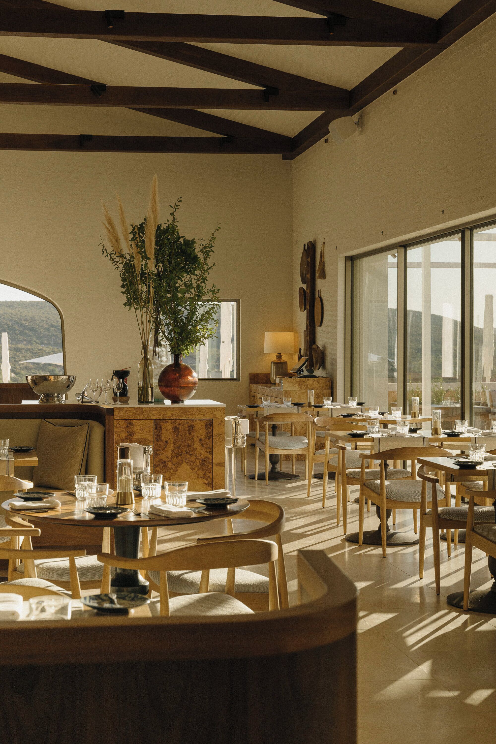 A dining area with multiple tables and chairs, a large window with a view, and a plant centerpiece.