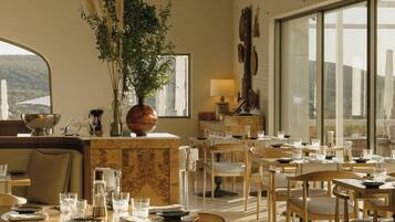 A dining area with multiple tables and chairs, a large window with a view, and a plant centerpiece.