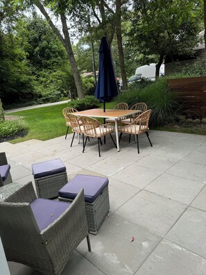 An outdoor patio with wicker furniture and a table, surrounded by greenery and trees.