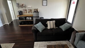 A living room with a dark wood floor, a black sofa with striped cushions, a coffee table with a woven coaster, and a kitchen area with a refrigerator and microwave. - Charming 4-bedroom house with AC and WiFi in amazing Cedar Hill (Cedar Hill)