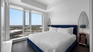 A bedroom with a large bed, a view of the city through floor-to-ceiling windows, and a nightstand with a lamp. - Midnight Rose-Your Elevated Stay! (Calgary)