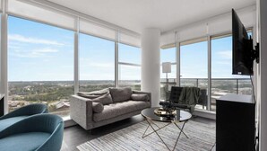 A spacious living room with a grey sofa, a blue armchair, and a coffee table with decorative items. Large windows offer a view of the cityscape. - Midnight Rose-Your Elevated Stay! (Calgary)