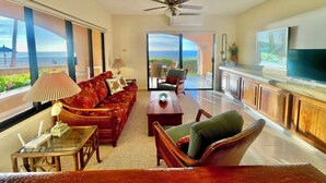 A spacious living room with a large sofa, a coffee table, and a TV. There is a ceiling fan and a view of the ocean through large windows.