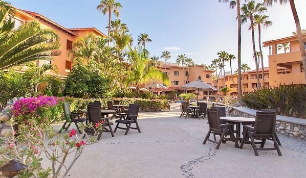 An outdoor patio area with multiple tables and chairs, surrounded by palm trees and a building with a tiled roof.