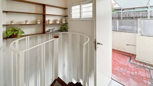 A room with a white curved bench, wooden shelves with jars, and a door leading to an outdoor area with a red-tiled floor.