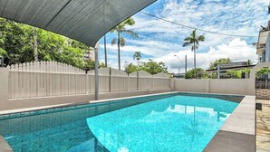 Une piscine rectangulaire à l’eau d’un bleu limpide, entourée d’une clôture blanche et bordée d’une zone ombragée.