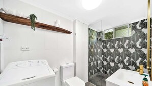 Une salle de bain moderne avec des toilettes blanches, un lave-linge, un lavabo avec un robinet doré et une douche avec un mur à motifs.