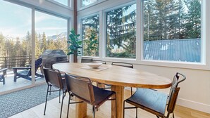 A dining area with a wooden table and chairs, large windows with a view of trees and mountains, and a barbecue grill outside. - Luxury Lodge w/ Breathtaking Mountain Views, large yard, fireplace  & WIFI (Snoqualmie Pass)