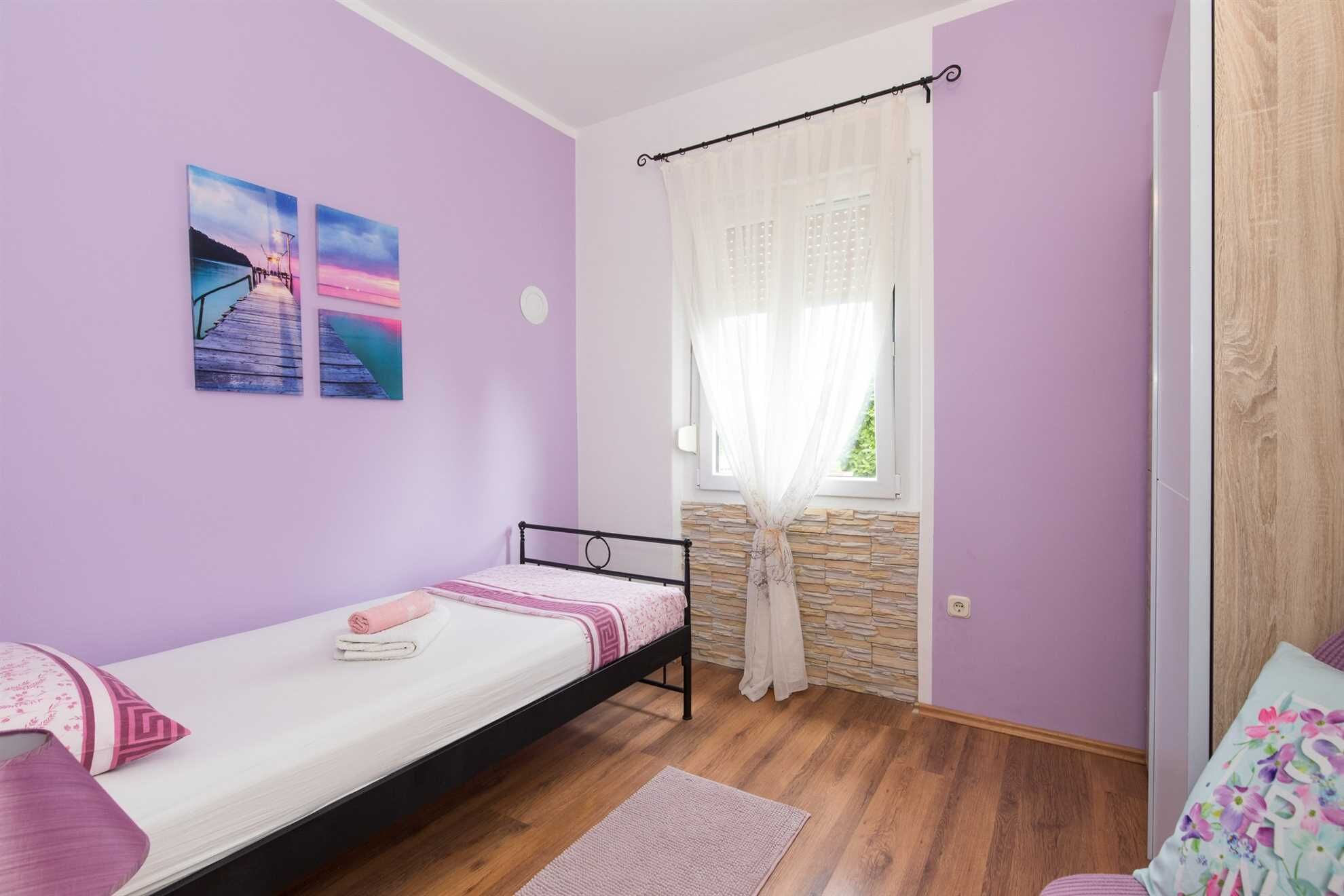 Un letto singolo con testiera, una finestra con tende leggere e due quadri appesi al muro.