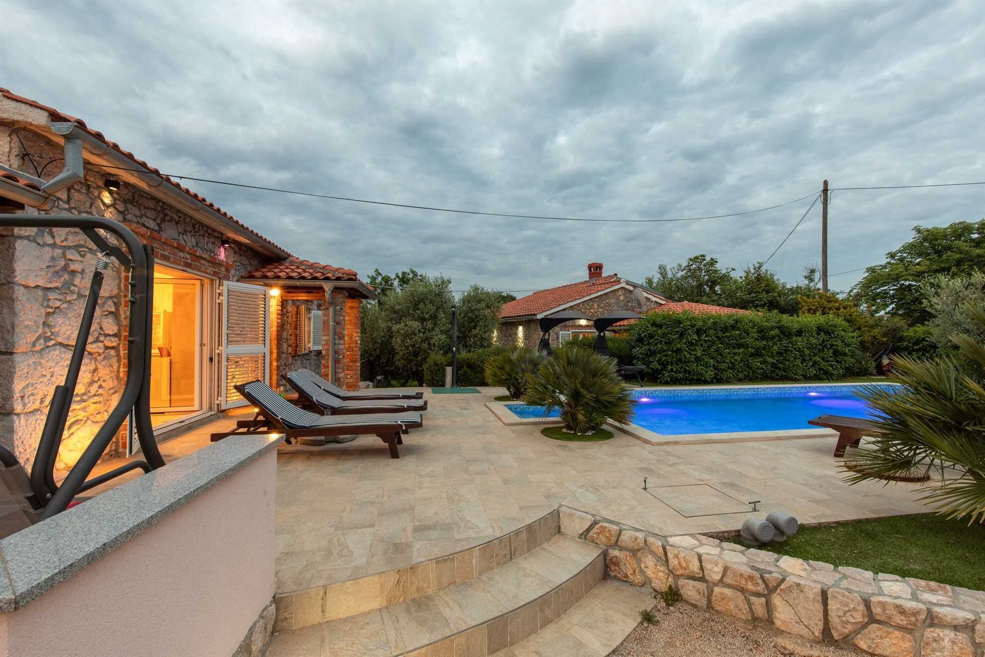 Ein Poolbereich mit Liegestühlen, einer Steinmauer und einer Steintreppe, die zu einer überdachten Terrasse führt.