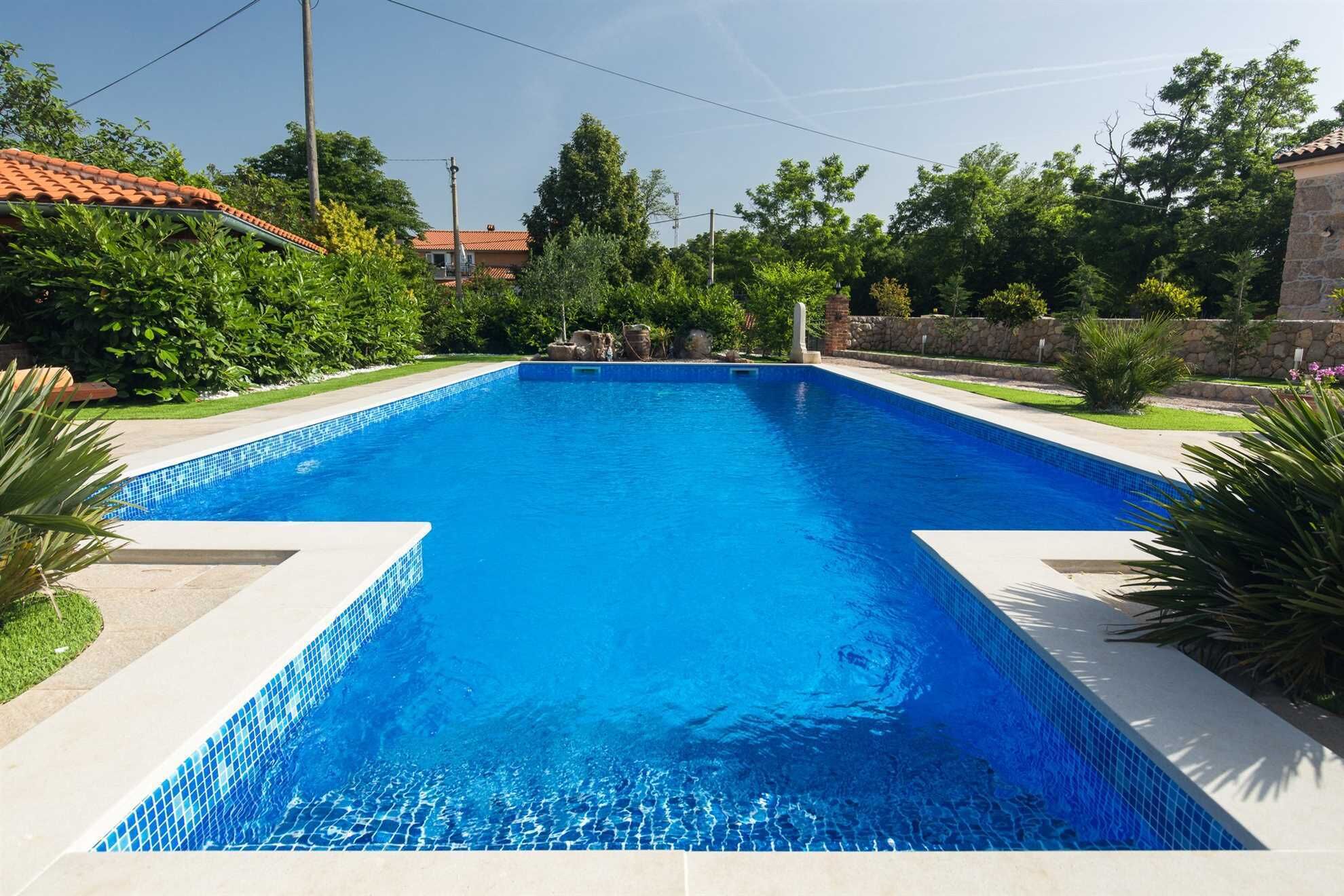 A rectangular swimming pool with clear blue water, surrounded by a stone wall and lush greenery.