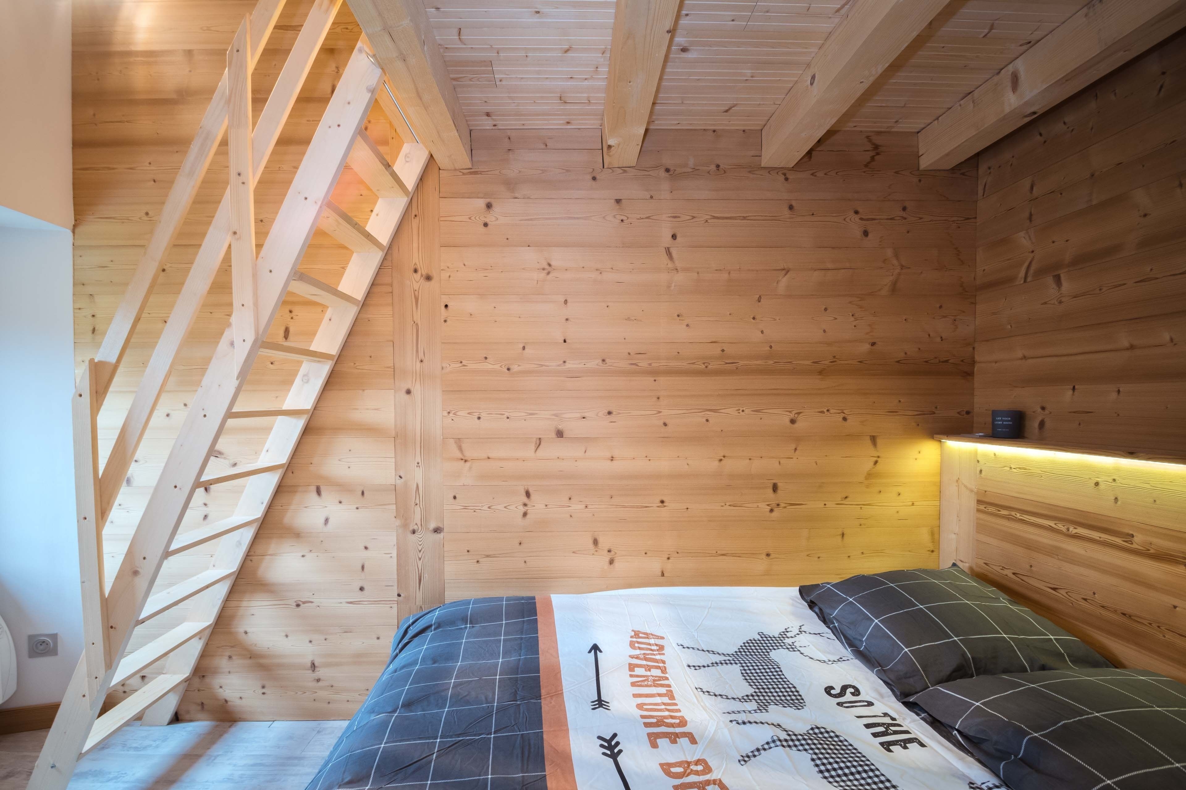 A wooden cabin room with a sloped ceiling, a bed with a 'Adventure Begins' themed bedspread, and a ladder leading to the upper sleeping area.