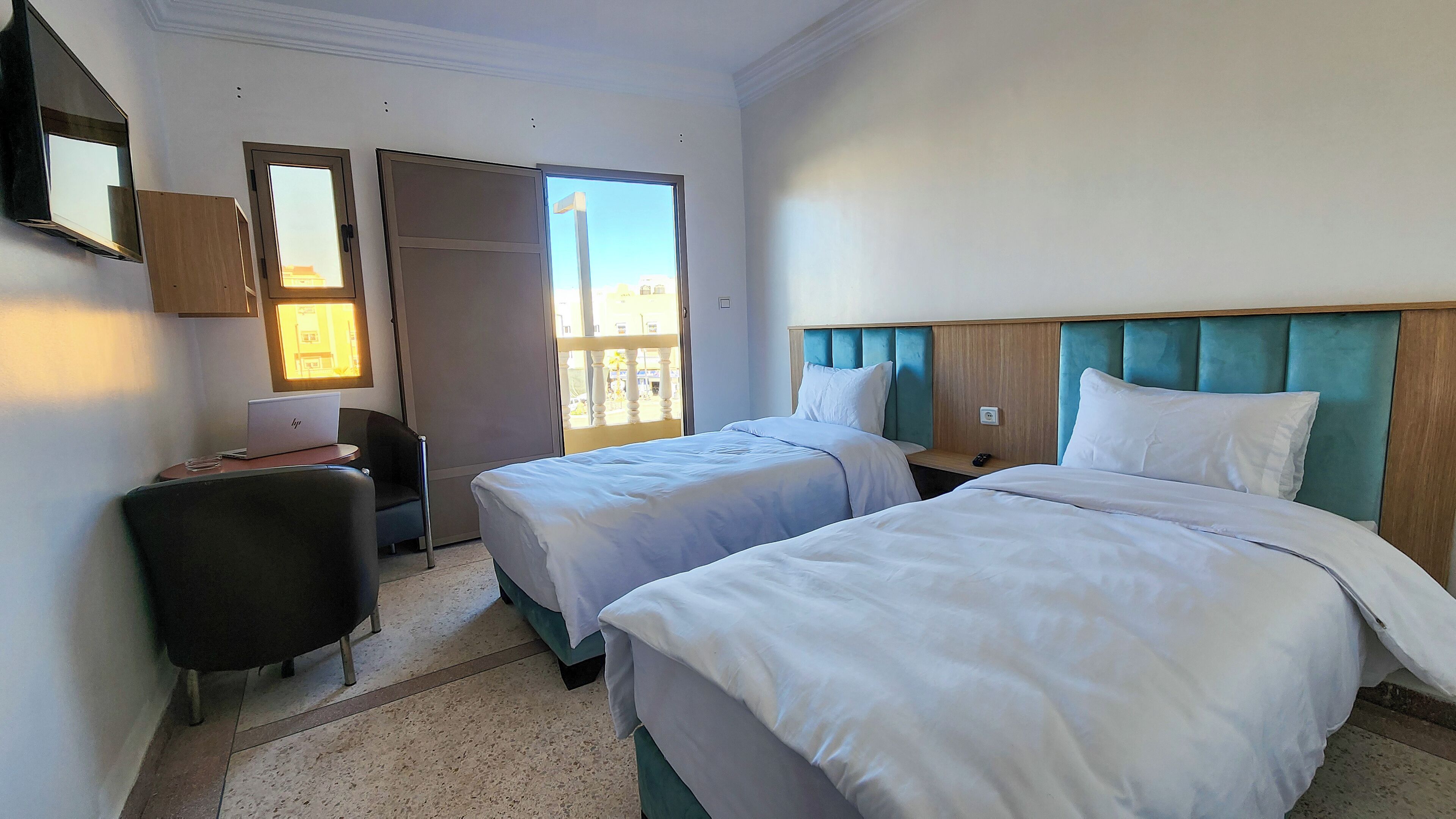 A hotel room with two beds, a desk with a laptop, a chair, and a window with a view of buildings.