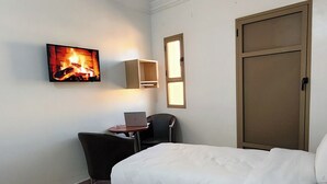 A hotel room with two beds, a desk with a laptop, a chair, a TV displaying a fireplace, and a door. - Hotel nakhil dak (Dakhla)