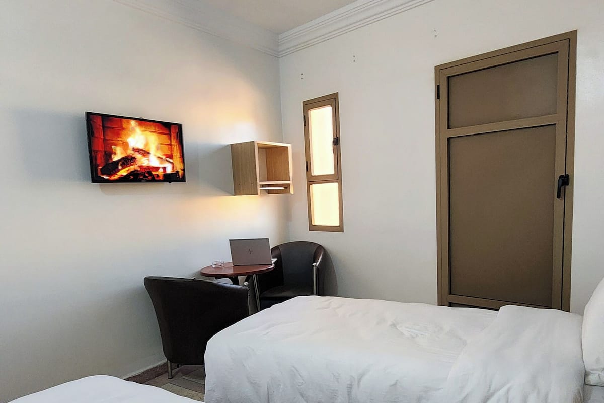 Une chambre d’hôtel avec deux lits, un bureau avec un ordinateur portable, une chaise, un téléviseur diffusant une image de cheminée et une porte.
