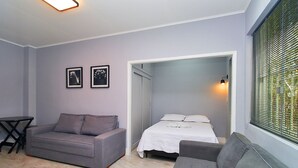 A compact living space with a grey sofa, a small table, a bed with white bedding, and two framed pictures on the wall. - Pompeu Loureiro (Rio de Janeiro)