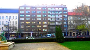 Ein mehrstöckiges Gebäude mit einer Reihe von Fenstern und Balkonen, ein grünes Schild für den Postdienst und eine Statue im Vordergrund.