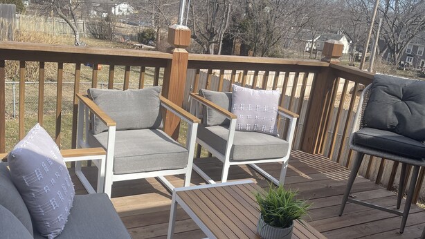 Una terraza de madera con un sofá y dos sillones, una mesita con una planta, y vistas a casas al fondo.