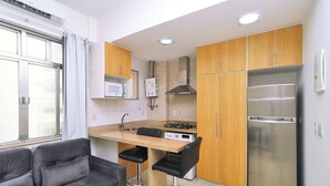 Cocina compacta con armarios de madera, un refrigerador de acero inoxidable, un microondas y una estufa a gas. Hay un taburete de bar y un sofá en el área de estar contigua.