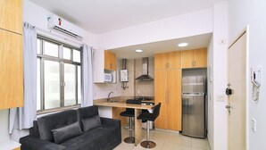 Una cocina compacta con refrigerador, microondas y estufa a gas. Hay un sofá negro y una pequeña mesa de comedor con dos sillas.