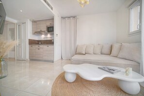 Uma sala de estar moderna com um sofá branco, uma mesa de centro branca e uma cozinha ao fundo.