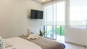 Un dormitorio moderno con una cama grande, un televisor de pantalla plana en la pared, una planta en maceta y vistas al verdor por la ventana.