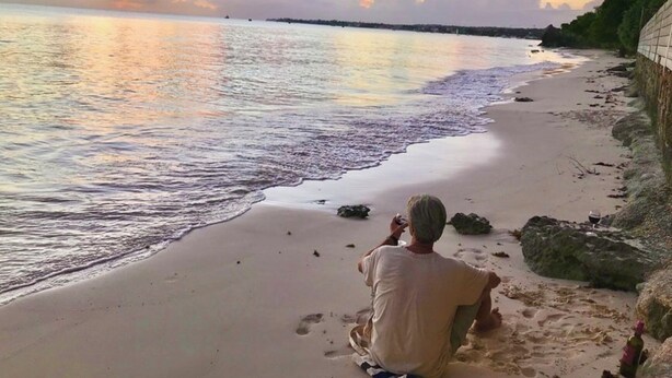 A man sitting on a sandy beach with a bottle and a glass, facing the ocean during sunset. - Villa Mia Apartment Studio #4 (Oistins)